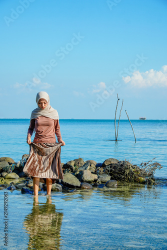 Cuadro en lienzo ​A young Asian Muslim woman in a hijab holding her skirt while standing in clear, shallow ocean water near a rocky shore under a bright blue sky
