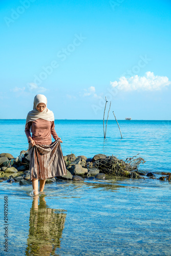 ​A young Asian Muslim woman in a hijab holding her skirt while standing in clear, shallow ocean water near a rocky shore under a bright blue sky. Concept of tropical vacation, modesty travel, peaceful