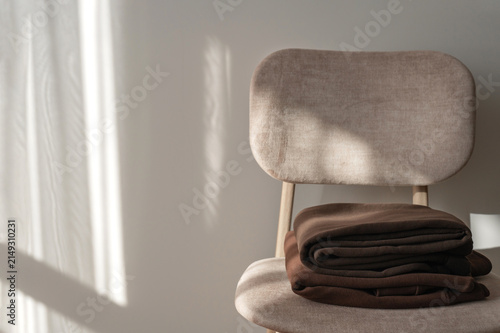 Brown jersey clothes on beige chair near empty beige wall background with abstract natural sunlight shadows, minimal cozy home interior still life, slow living