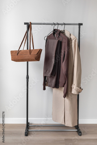 Minimal aesthetic fall fashion capsule wardrobe. Clothing rack with brown clothes, beige trench coat and caramel brown sueded bag, neutral beige empty room interior background