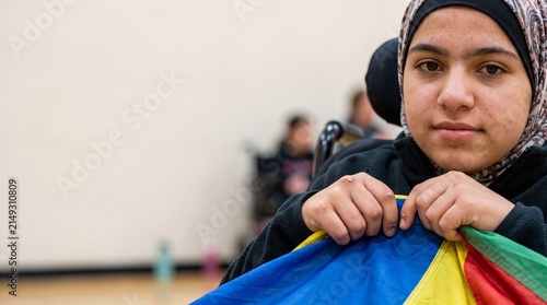 Young Muslim woman in wheelchair holding colorful parachute, looking at camera with determined expression