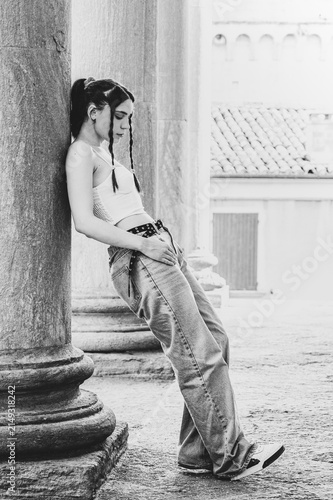 Monochrome black and white full body portrait of a trendy young woman with braided hair leaning against an ancient column looking in profile outdoors