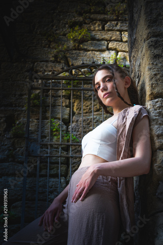 Dramatic low angle portrait of a cool young woman with braided hair leaning against a rough stone wall looking away into light near a historic iron gate