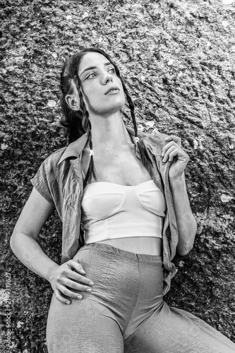 Monochrome black and white fashion portrait of a stylish young female model with braids wearing casual earth tone clothing posing against a natural rough stone wall
