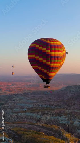 Vertical video. Goreme, Nevsehir, Turkey. A single large checkered hot air balloon in vibrant yellow, red, orange and purple, drifting over the eroded volcanic landscape of Cappadocia at sunrise, aer