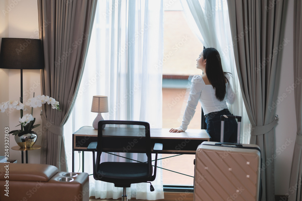 Obraz premium Young woman enjoying hotel room view, standing by window with curtains, feeling relaxed during business travel or vacation stay in modern accommodation, arriving with suitcase