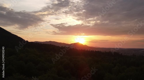Aerial view cinematic shot of mountains and amazing sunset or sunrise in summer time. Silhouette of green forest slopes of mountain range. Bright disk of sun in sky slowly sets tops of mountains