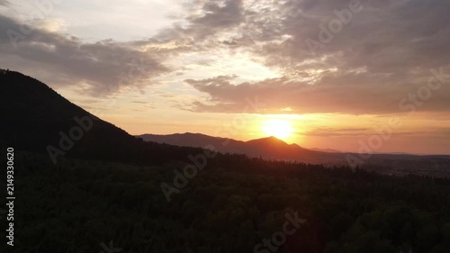 Aerial drone view of beautiful mountains at warm sunset. Forest slopes of mountain range. Bright disk of sun slowly sets tops of mountains. Dramatic sunrise sky background