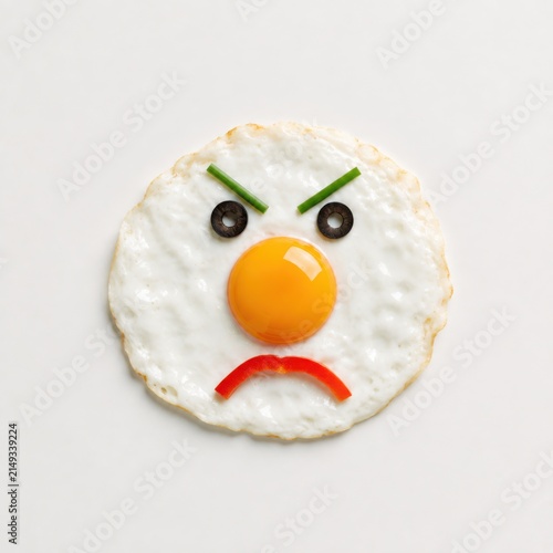 Angry fried egg face made with vegetables on white background