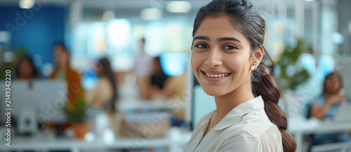 a smiling youthful indian employee, working on a large screen monitor, brightly lit office space