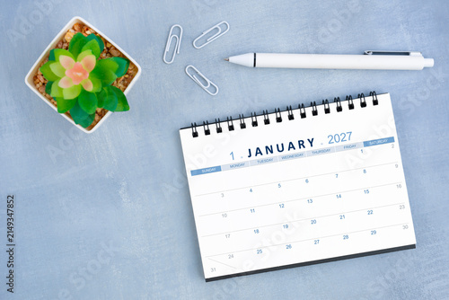 January 2027 desk calendar for 2026 and plant pot with pen on blue cement, Flat lay.