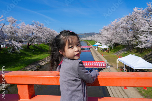 Little Asian Girl on Red Bridge Looking Back with Cherry Blossoms and River Background in Japan. Asian girl standing on a vermilion Japanese bridge. cherry blossom Sakura festival along a riverbank.
