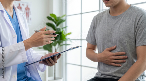 Doctor consulting male patient with stomach pain in clinic, reviewing notes on clipboard during examination