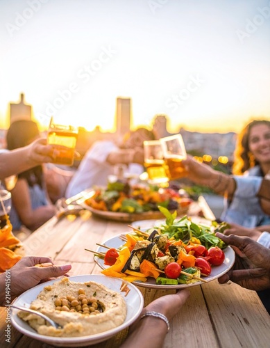 Diverse friends enjoy a delicious outdoor rooftop dinner party at golden hour sunset, passing healthy food and toasting drinks, celebrating joy, friendship, and summer lifestyle.