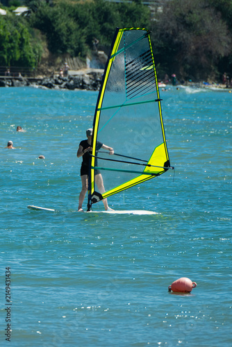 Quadro su tela A person enjoys a sunny summer day windsurfing on sparkling blue water with a vibrant yellow and transparent sail near a lush coastline