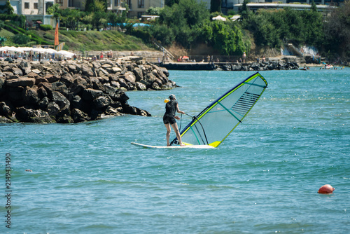 Foto A windsurfer balances on their board while pulling up a yellow and transparent sail in bright blue water near a rocky, tree-lined coastal resort