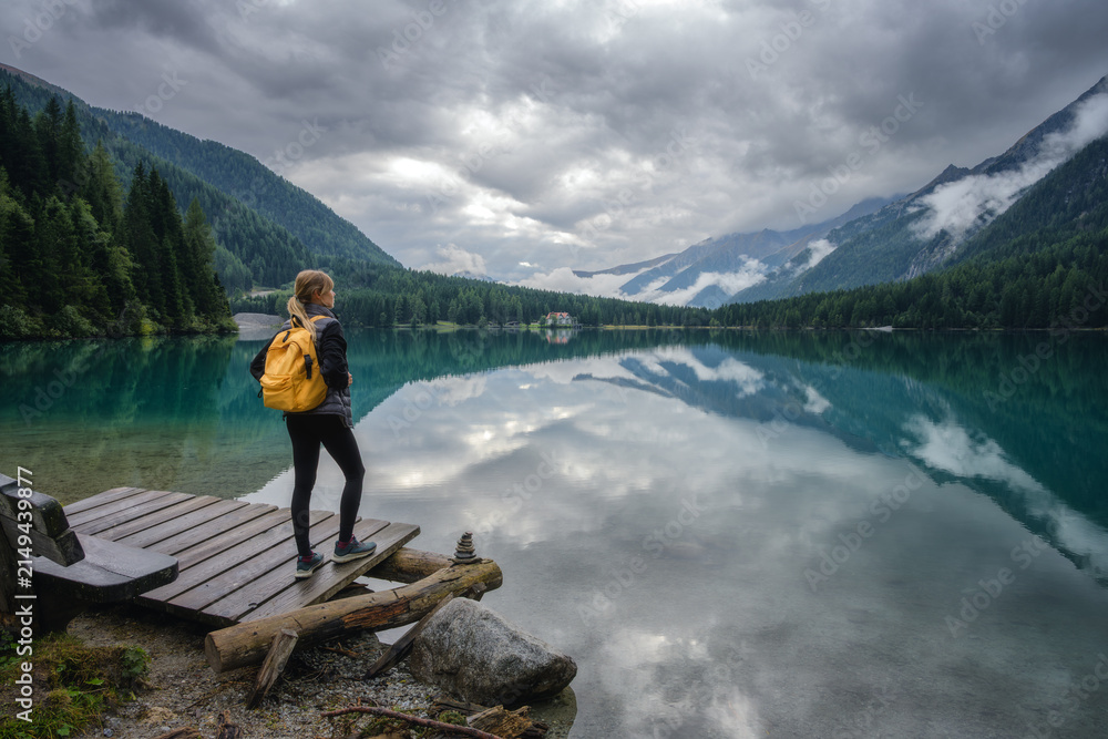 Naklejka premium Woman hiker with yellow backpack standing on wooden pier at alpine lake in Dolomites, Italy. Mountain landscape reflected in calm water with forest and cloudy sky on overcast summer rainy day. Travel