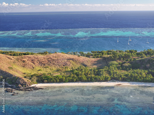 Aerial view of Mana Island featuring a pristine white sand beach, lush green hills, and a vibrant turquoise lagoon with coral reefs and sailboats in Western Division, Fiji.