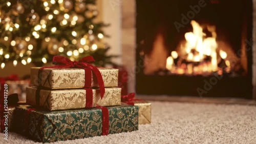 Cosy festive scene featuring wrapped Christmas presents resting on soft carpet before bright burning fireplace with decorated evergreen tree in blurred background.