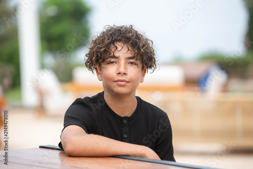 Thoughtful curly-haired teenage boy sitting outdoors and looking at camera with calm confident expression. Portrait of a curly-haired teenage boy sitting outdoors at a table 