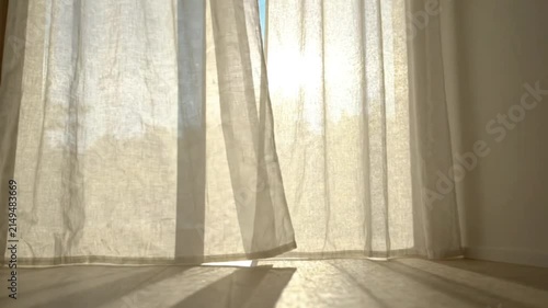 Sunlight creating beautiful light and shadow patterns on floor as white curtains gently sway with the breeze in a bright room with window view of green trees.