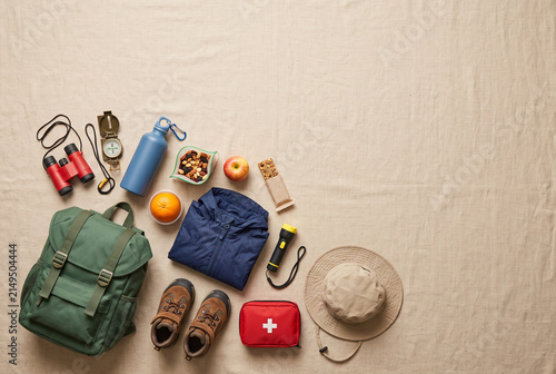 A range of hiking supplies is laid out on a light surface, including a backpack, water bottle, flashlight, snacks, and clothing. Perfect for a day spent in nature