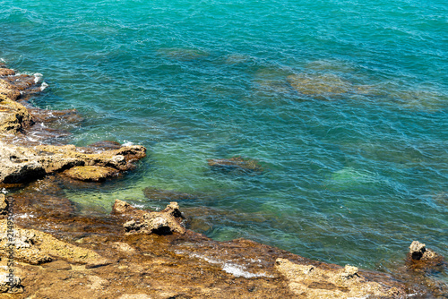Turquoise sea water lapping over rocky coastal shoreline on sunny day, clear ocean waves with visible underwater rock formations and vibrant teal color