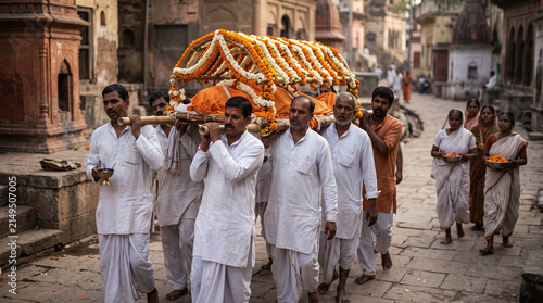 A traditional funeral procession walking along a stone-paved street in an ancient town, with a group of men dressed in white carrying a bamboo bier adorned with orange and white marigold flowers