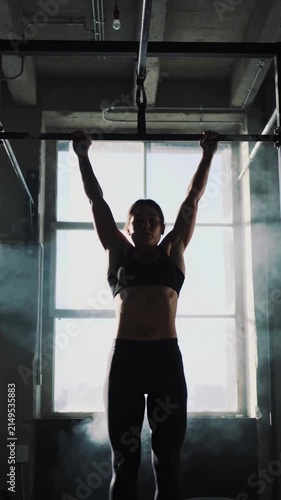 Athletic woman doing pull-ups in dark gym with bright window background and smoke, medium shot
