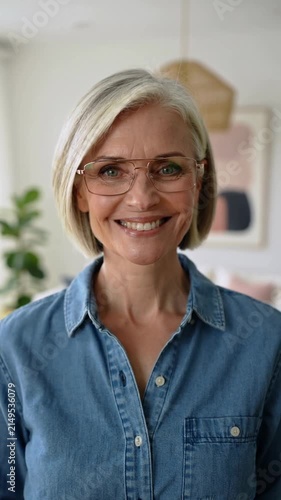 Mature woman with blonde hair wearing blue denim shirt and glasses looking at camera, one shot