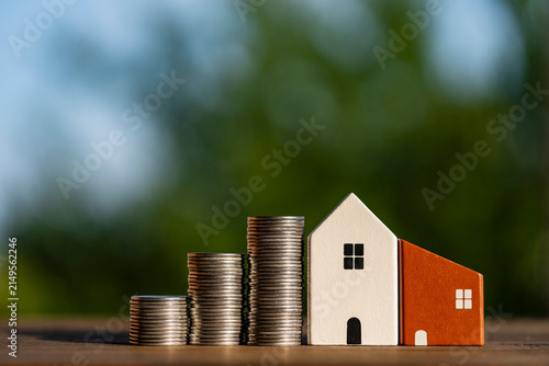Wooden toy houses next to stacks of coins outdoors. Closeup view of small wooden toy houses standing in line next to growing stacks of silver coins on wooden surface against green nature background