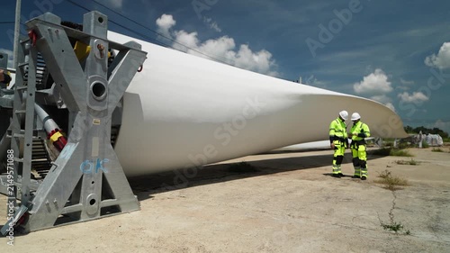 Workers inspect a large wind turbine blade in a manufacturing area during the day