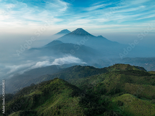 Aerial view of Gunung Prau surrounded by lush green hills, rolling clouds, and mist under a clear blue sky in Bawang, Central Java, Indonesia.