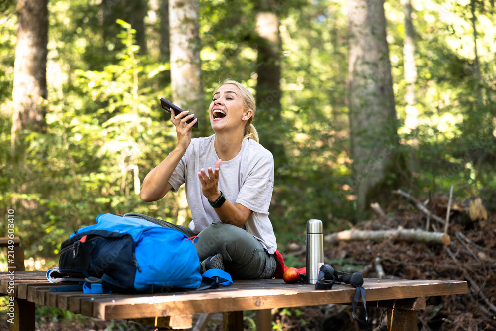 Fototapeta premium Happy woman sitting on a wooden bench, laughing while talking on her phone during a hiking trip in the green forest