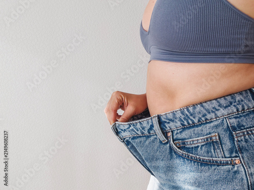 Woman showing loose jeans around waist, cropped torso in blue top, visible gap between body and denim, weight loss, slimming, fitness, healthy lifestyle, body transformation concept