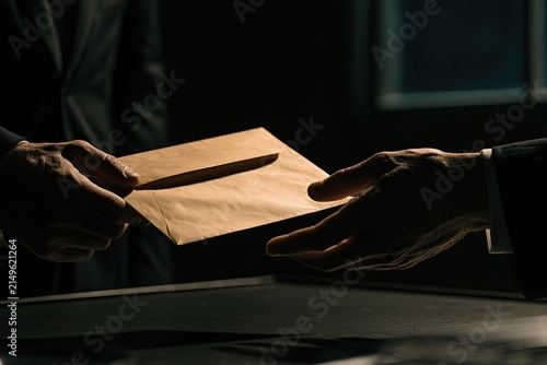 Hands exchange a brown envelope in a shadowy, clandestine meeting