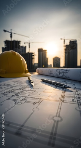 Obraz na płótnie Yellow Safety Helmet and Architectural Blueprints with High Rise Construction at Sunset, Contractor Engineering, Construction Planning, Architecture and Urban Development Concept