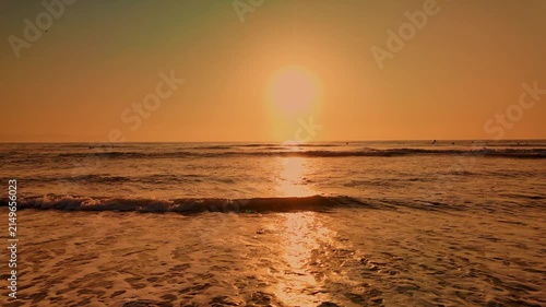 Low angle perspective capturing gentle waves rolling onto a wet sandy shore as the sun sets. Warm golden light reflects off the rippled water surface and glistening sand textures.