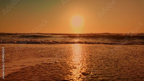 Low angle perspective capturing gentle waves rolling onto a wet sandy shore as the sun sets. Warm golden light reflects off the rippled water surface and glistening sand textures, creating a serene mo