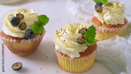 Fresh vanilla cupcakes featuring rich white cream frosting, blueberries, and mint are artfully arranged on a textured surface under bright natural light