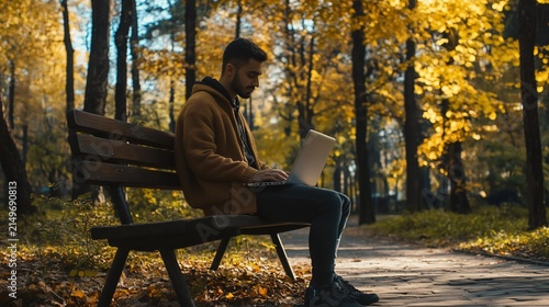 Young man working on a laptop while sitting on a bench in autumn park
