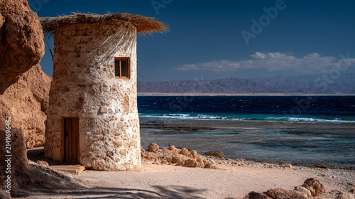 Rustic stone structure with thatch roof situated on a sandy beach by the clear blue sea