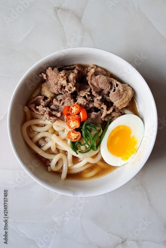 A bowl of udon noodles in a savory broth, topped with green onions, tempura, and a soft boiled egg. The thick, chewy noodles and rich flavors make it a comforting Japanese classic.