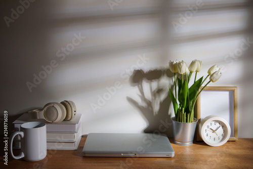 Cozy home workspace with a closed laptop, headphones, books, coffee mug, flowers, and clock arranged on a wooden desk.
