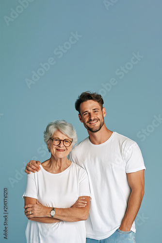Young Bearded Man with Elderly Woman, Mother and Son, Affectionate Intergenerational Bond and Family Connection, Smiling Together in White T-Shirts Against Blue Studio Background. Copy Space, 2:3