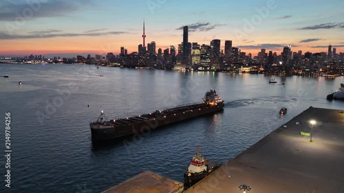 High-angle stationary shot looking down at a large bulk carrier vessel navigating past a concrete industrial pier, revealing the illuminated downtown Toronto skyline and CN Tower at dusk