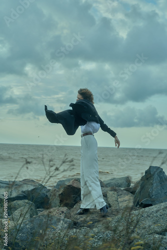 Bild auf Leinwand Wind swept figure on rocky coast, flowing coat and white pants, ocean horizon in the background, outdoor fashion and freedom in motion, dramatic sea breeze and bold stance