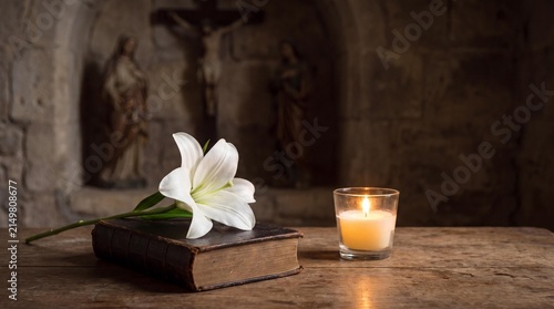 Sacred Serenity: A white lily and a lit candle rest upon an antique book, set against a backdrop of religious statuary, evoking solemn contemplation and spiritual peace.