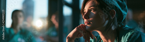 Healthcare professional in thoughtful break beside window, nurse in surgical cap with reflective gaze and soft light