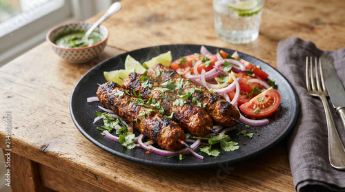 Grilled meat kebab skewers served on dark ceramic plate with fresh tomato salad sliced red onion cilantro and lime wedges on rustic wood table.